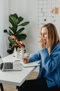 A woman sitting at her desk with a plant in the background. She is working at her computer and sipping a glass of water.